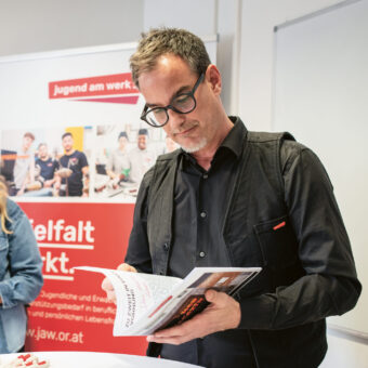 Ein Mann liest an einem runden Tisch in einer Broschüre, während drei Frauen daneben stehen; im Hintergrund ist ein rot-weißes Veranstaltungsbanner zu sehen.