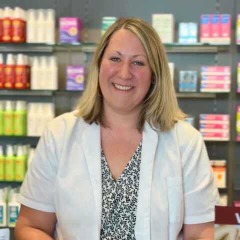 Eine Frau in einem weißen Kittel lächelt vor den Regalen einer Apotheke, die mit verschiedenen Produkten und Medikamentenpackungen gefüllt sind.