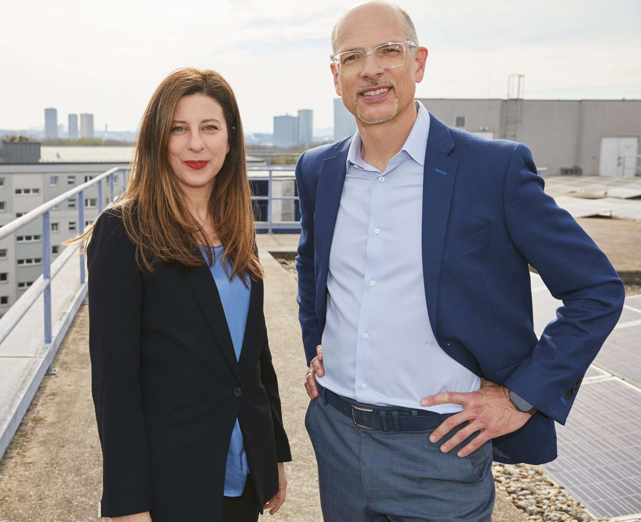 Zwei Personen in Geschäftskleidung stehen auf einem Dach, im Hintergrund sind Gebäude und die Skyline der Stadt zu sehen, und lächeln in die Kamera.