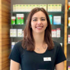Eine Frau mit glattem braunem Haar und einem schwarzen Shirt lächelt in einer Apotheke, hinter ihr stehen Regale mit Medikamenten und anderen Produkten.