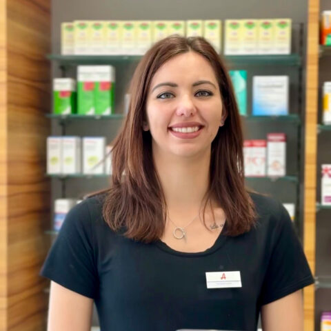 Eine Frau mit glattem braunem Haar und einem schwarzen Shirt lächelt in einer Apotheke, hinter ihr stehen Regale mit Medikamenten und anderen Produkten.