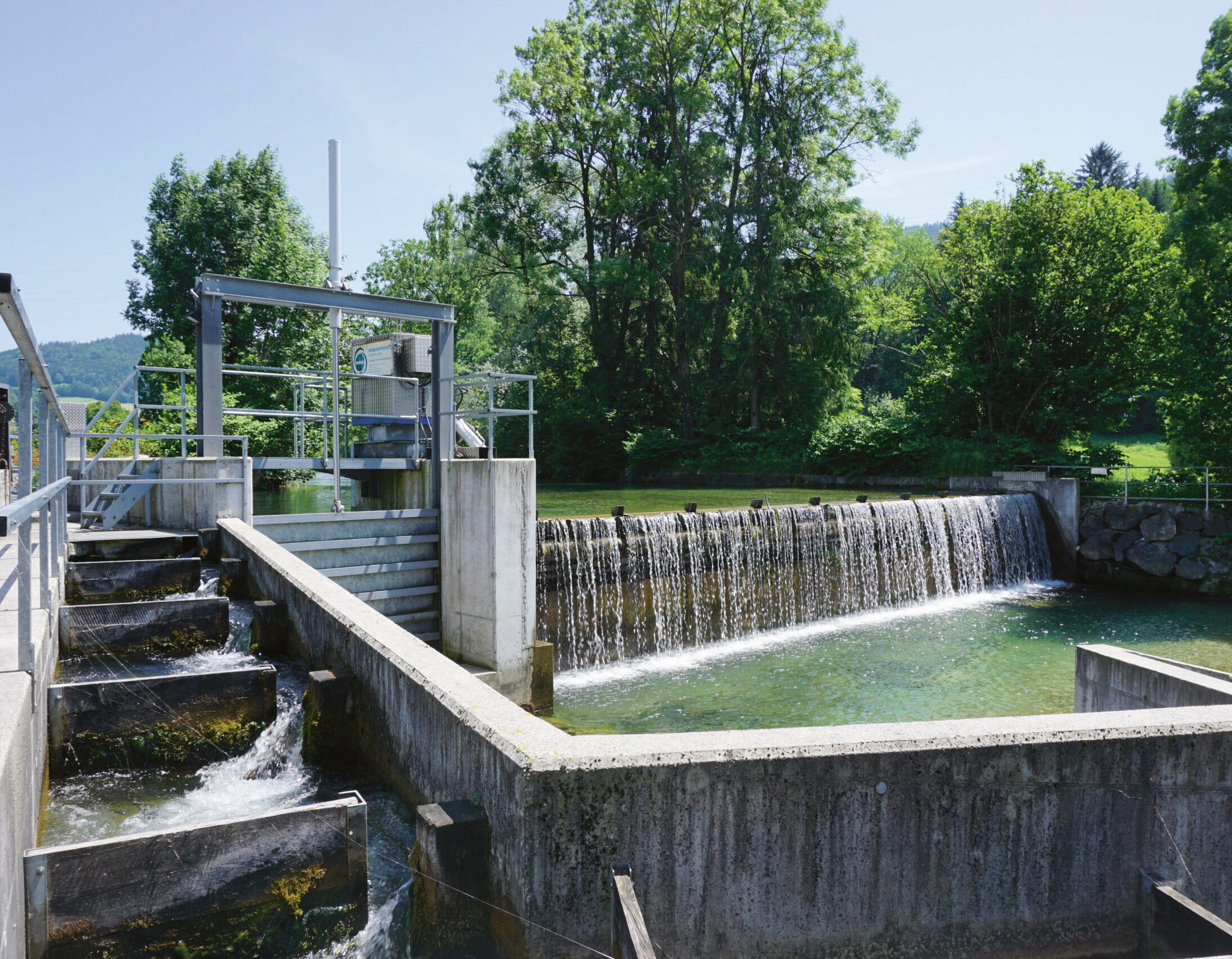 Ein kleiner Wasserkraftdamm mit einer Fischtreppe an einem sonnigen Tag, umgeben von grünen Bäumen und klarem Wasser, das über das Bauwerk fließt.