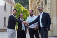 Vier Menschen lächeln und reichen sich auf einer Kopfsteinpflasterstraße die Hände, während sie vor historischen Gebäuden ihren Erfolg bei den WKO Steiermark Forschungsstipendien 2026 feiern.