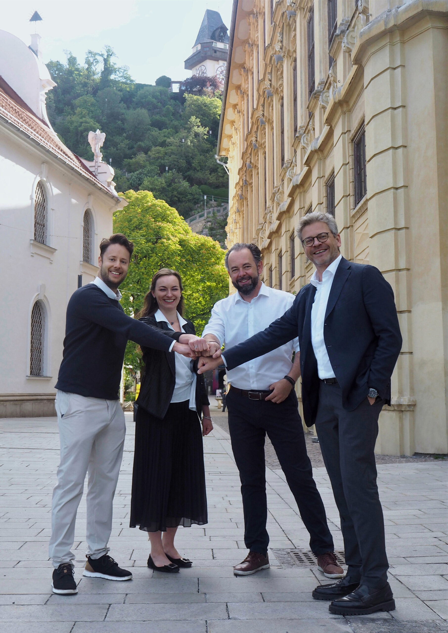 Vier Menschen lächeln und reichen sich auf einer Kopfsteinpflasterstraße die Hände, während sie vor historischen Gebäuden ihren Erfolg bei den WKO Steiermark Forschungsstipendien 2026 feiern.