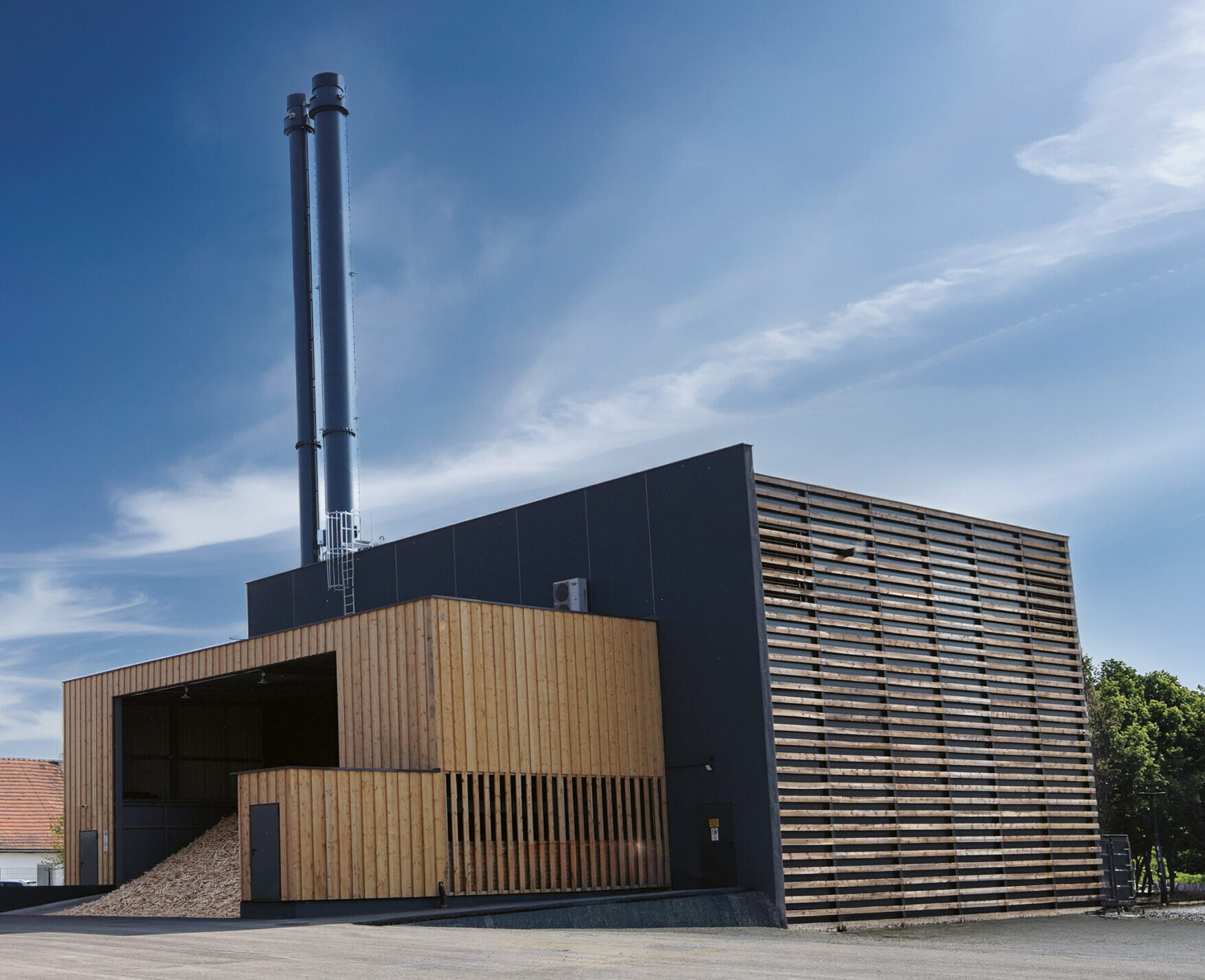 Modernes Industriegebäude mit Holzverkleidung, hohen Schornsteinen und einem großen Haufen Holzpellets neben einem Schild mit der Aufschrift „Steirer Pellets“ unter blauem Himmel.