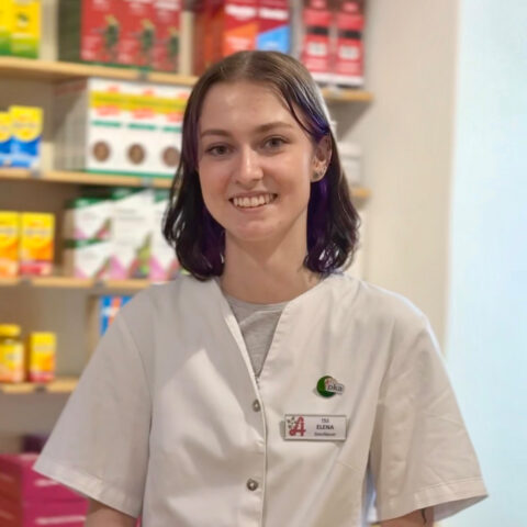 Eine Frau in einem weißen Apothekenkittel steht lächelnd vor Regalen, die mit bunten Medikamentenschachteln gefüllt sind.