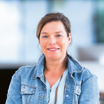 Eine Frau in einer Jeansjacke und einem gestreiften Kleid steht mit verschränkten Armen lächelnd in einem modernen Büro, im Hintergrund sind verschwommene Computerbildschirme zu sehen.