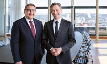 Zwei Männer in Anzügen stehen lächelnd in einem modernen Konferenzraum mit einem langen Tisch und Blick auf die Stadt durch große Fenster.