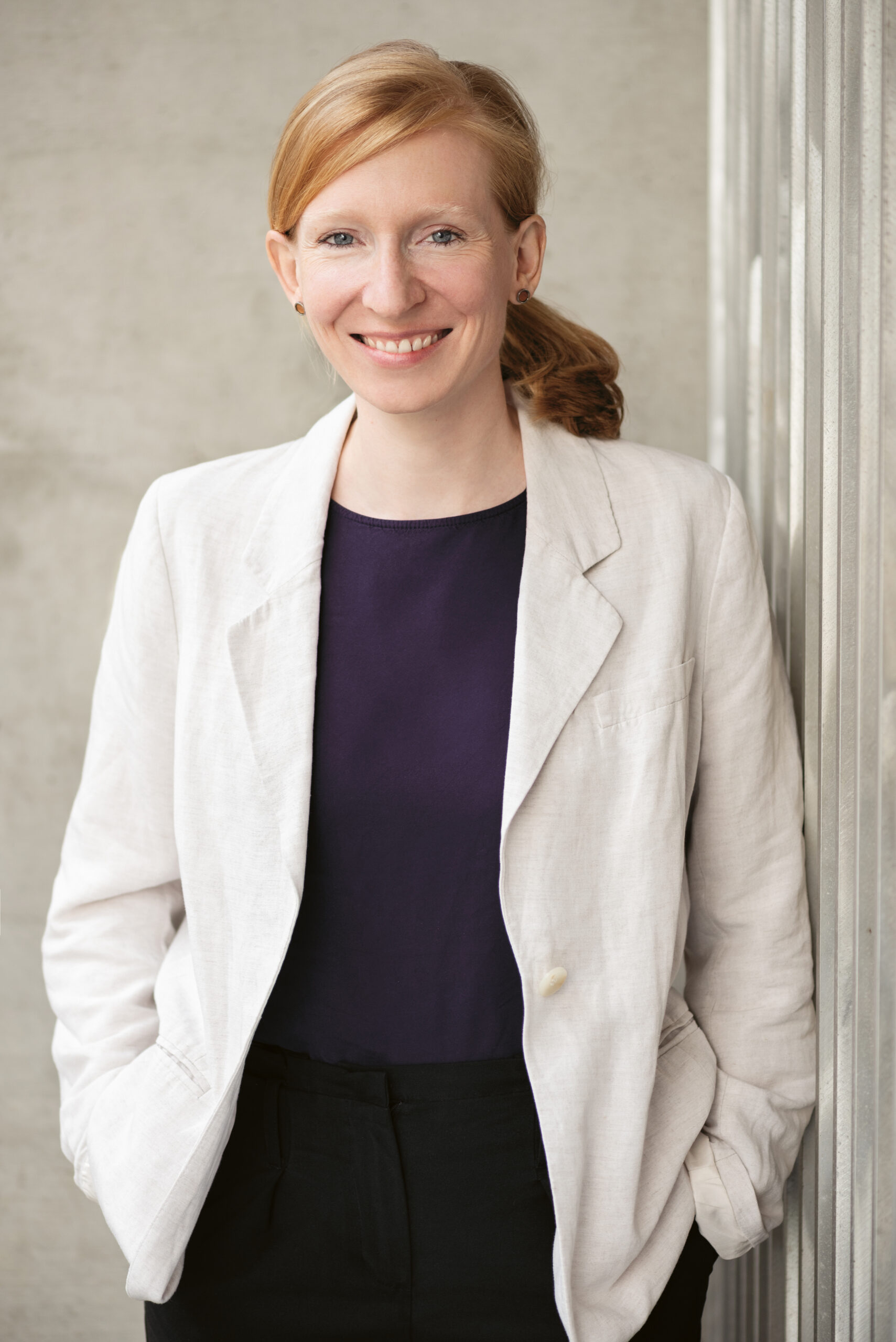 Eine Frau mit roten Haaren, die zu einem Pferdeschwanz zusammengebunden sind, trägt einen weißen Blazer und ein dunkles Hemd und steht lächelnd mit den Händen in den Taschen vor einer hellen Steinmauer.