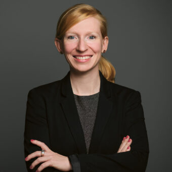 Eine Frau mit blonden Haaren, die zu einem Pferdeschwanz zusammengebunden sind, trägt einen schwarzen Blazer und lächelt mit verschränkten Armen vor einem schlichten dunkelgrauen Hintergrund.