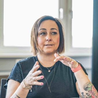Eine Frau mit Tattoos gestikuliert, während sie in einem Büro spricht; sie sitzt auf einem Stuhl, im Hintergrund ist ein Fenster zu sehen.