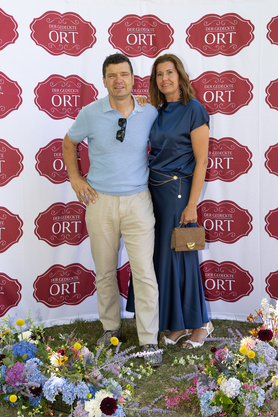 Ein Mann und eine Frau posieren gemeinsam vor einem „Der gedeckte Ort, Band 2“-Banner und bunten Blumen bei einer Veranstaltung im Freien.