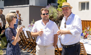 Drei Erwachsene in Sommerkleidung unterhalten sich mit einem Getränk in der Hand an einem langen Tisch – eine sonnendurchflutete Szene eines Essens im Freien für „Der gedeckte Ort, Band 2“.