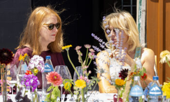 Zwei Frauen mit Sonnenbrillen unterhalten sich an einem sonnigen Tisch, umgeben von Blumen und Wasserflaschen – „Der gedeckte Ort, Band 2“ bildet den Rahmen für ihren Tag.