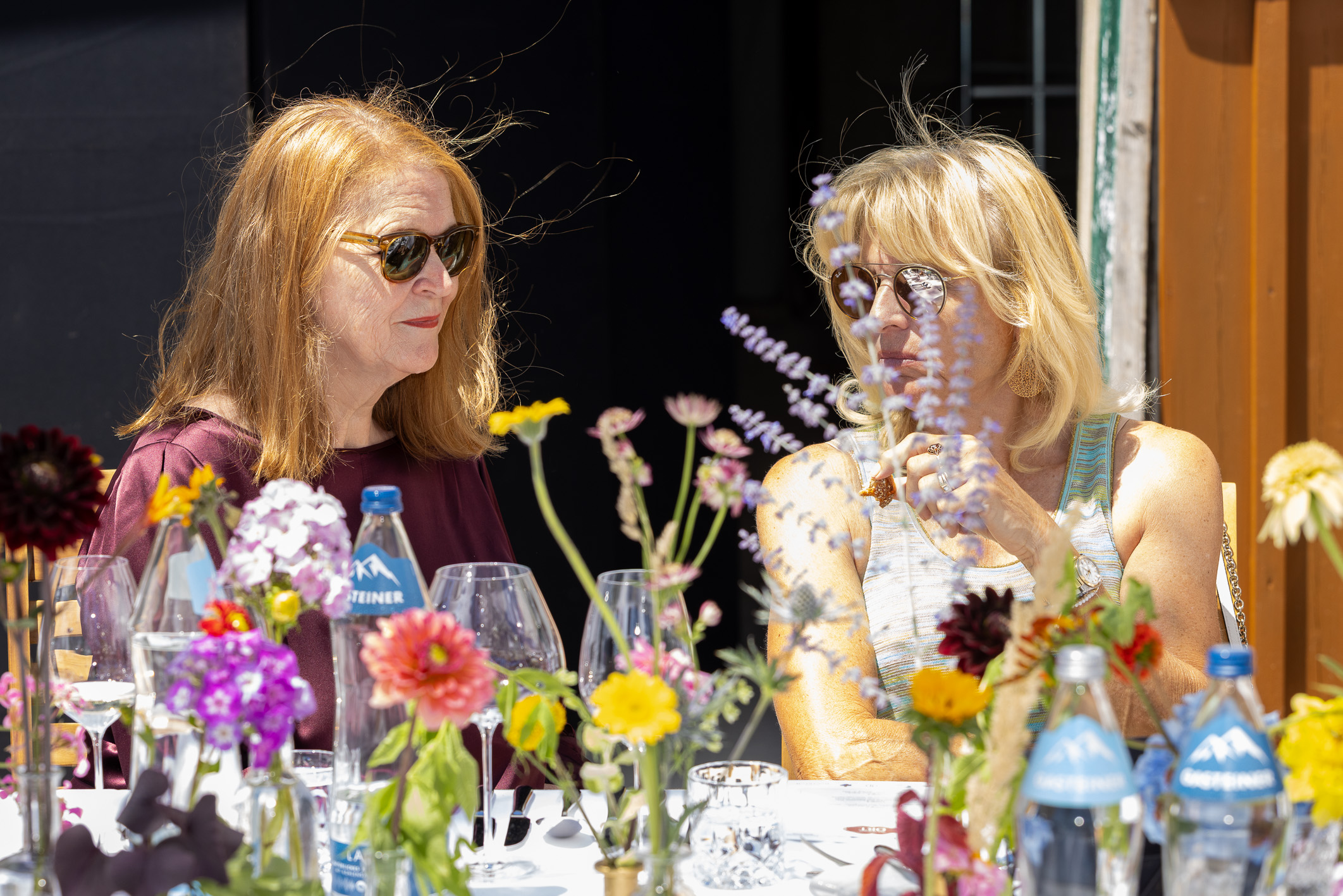 Zwei Frauen mit Sonnenbrillen unterhalten sich an einem sonnigen Tisch, umgeben von Blumen und Wasserflaschen – „Der gedeckte Ort, Band 2“ bildet den Rahmen für ihren Tag.
