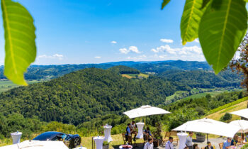 Eine große Gruppe speist im „Der gedeckte Ort, Band 2“ im Freien an einem langen Tisch mit Blick auf die Hügel, umgeben von üppigem Grün und unter weißen Sonnenschirmen vor strahlend blauem Himmel.
