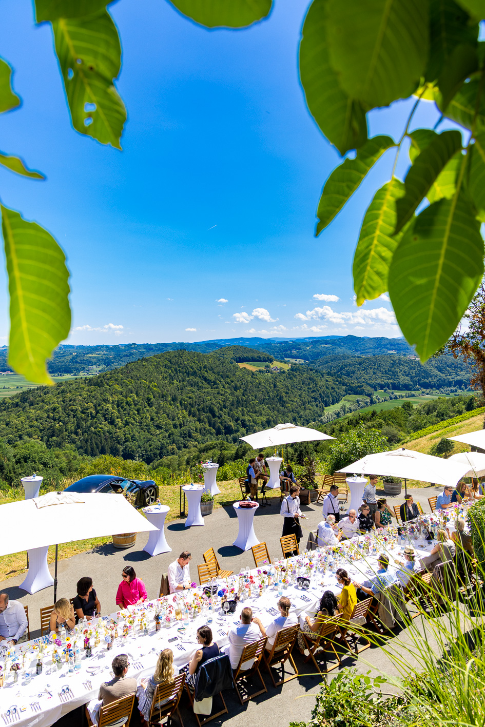 Eine große Gruppe speist im „Der gedeckte Ort, Band 2“ im Freien an einem langen Tisch mit Blick auf die Hügel, umgeben von üppigem Grün und unter weißen Sonnenschirmen vor strahlend blauem Himmel.