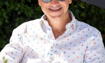 Ein Mann mit Sonnenbrille und Strohhut lächelt im Freien in einem weiß gemusterten Hemd, umgeben von Grünpflanzen und Blumen – und fängt damit die Atmosphäre von „Der gedeckte Ort, Band 2“ ein.