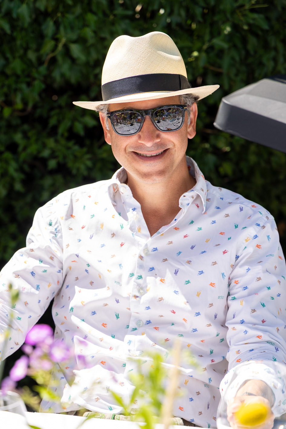 Ein Mann mit Sonnenbrille und Strohhut lächelt im Freien in einem weiß gemusterten Hemd, umgeben von Grünpflanzen und Blumen – und fängt damit die Atmosphäre von „Der gedeckte Ort, Band 2“ ein.
