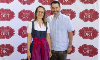 Eine lächelnde Frau in bayerischer Tracht und ein Mann in Freizeitkleidung posieren vor einer Veranstaltungskulisse von „Der gedeckte Ort“.