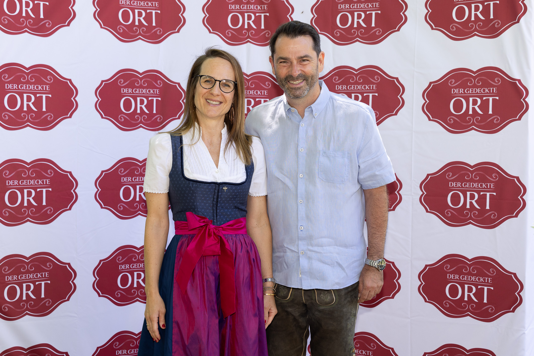 Eine lächelnde Frau in bayerischer Tracht und ein Mann in Freizeitkleidung posieren vor einer Veranstaltungskulisse von „Der gedeckte Ort“.