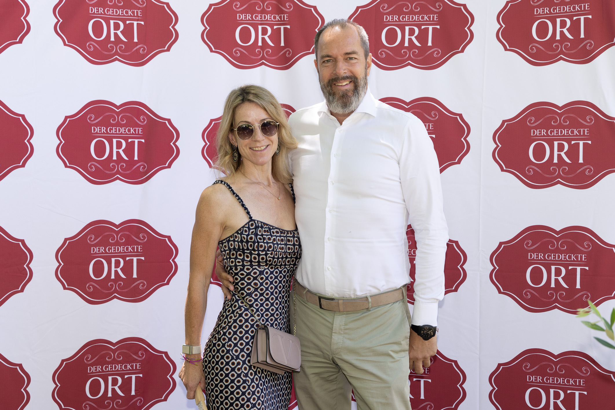 Ein lächelndes Paar posiert vor einem Hintergrund mit roten „Der Gedeckte Ort“-Logos; die Frau trägt ein gemustertes Kleid, der Mann ein weißes Hemd.