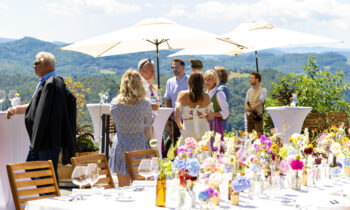 Bei einer Veranstaltung im Freien, vor dem Hintergrund von Bergen und grüner Natur, mischen sich Menschen in Sommerkleidung zwischen hohen Tischen und Blumenarrangements.