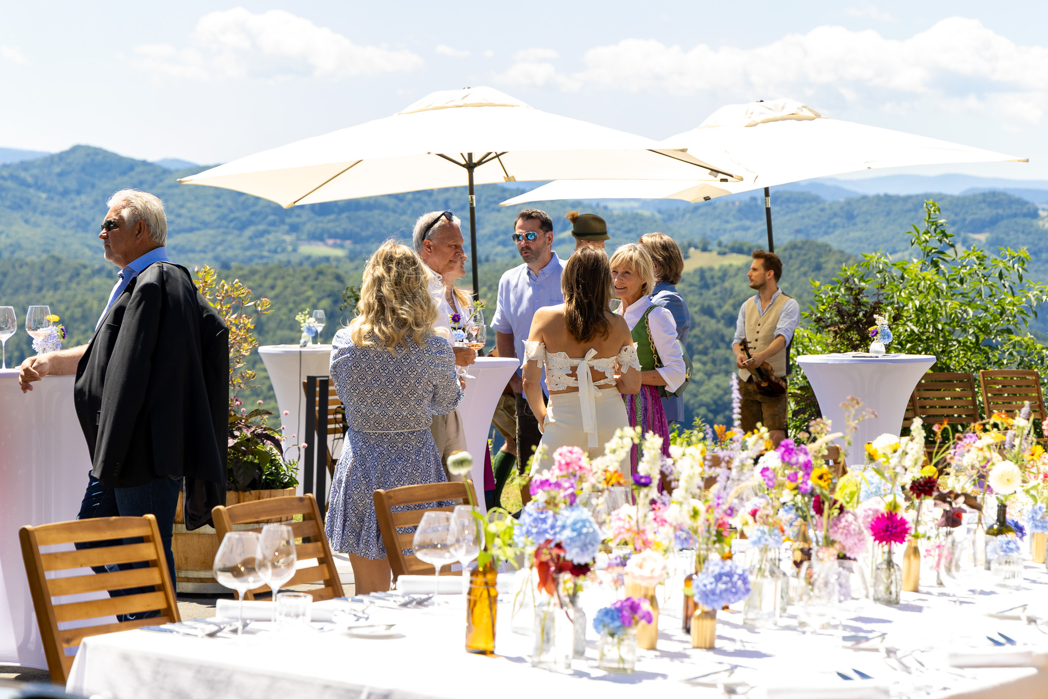 Bei einer Veranstaltung im Freien, vor dem Hintergrund von Bergen und grüner Natur, mischen sich Menschen in Sommerkleidung zwischen hohen Tischen und Blumenarrangements.