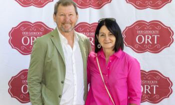 Ein Mann in einer hellgrünen Jacke und eine Frau in einer leuchtend rosa Bluse lächeln vor einem „Der Gedeckte Ort“-Banner mit Wiederholungsmuster.