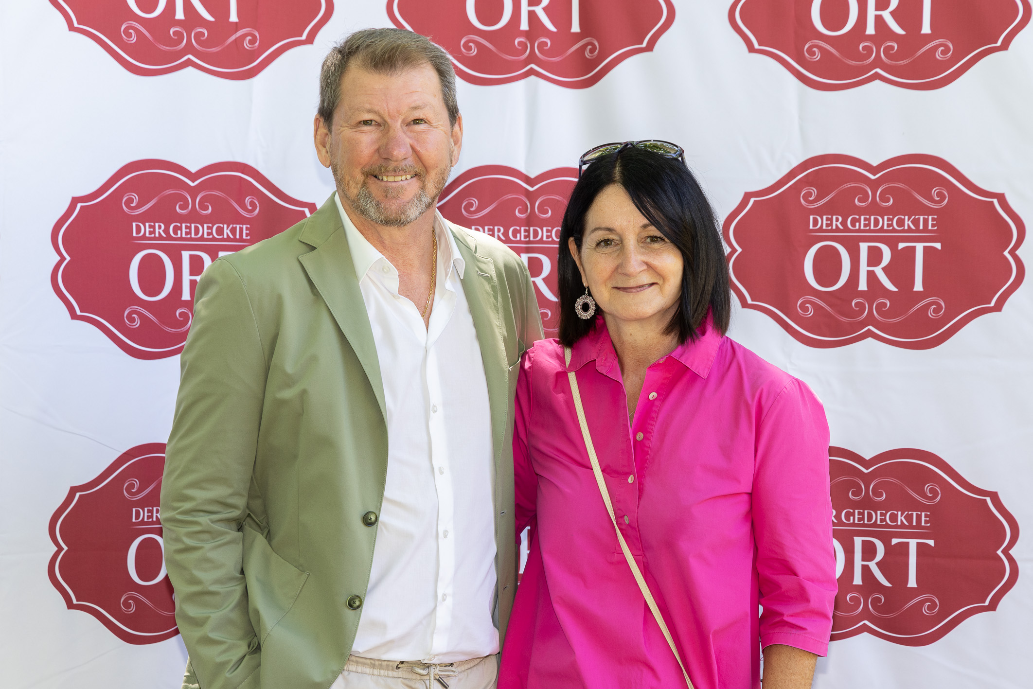 Ein Mann in einer hellgrünen Jacke und eine Frau in einer leuchtend rosa Bluse lächeln vor einem „Der Gedeckte Ort“-Banner mit Wiederholungsmuster.