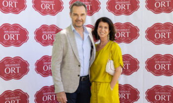 Ein Mann in einem grauen Blazer und eine Frau in einem gelben Kleid stehen lächelnd vor einer rot-weißen Veranstaltungskulisse mit der Aufschrift „Der gedeckte Ort“.