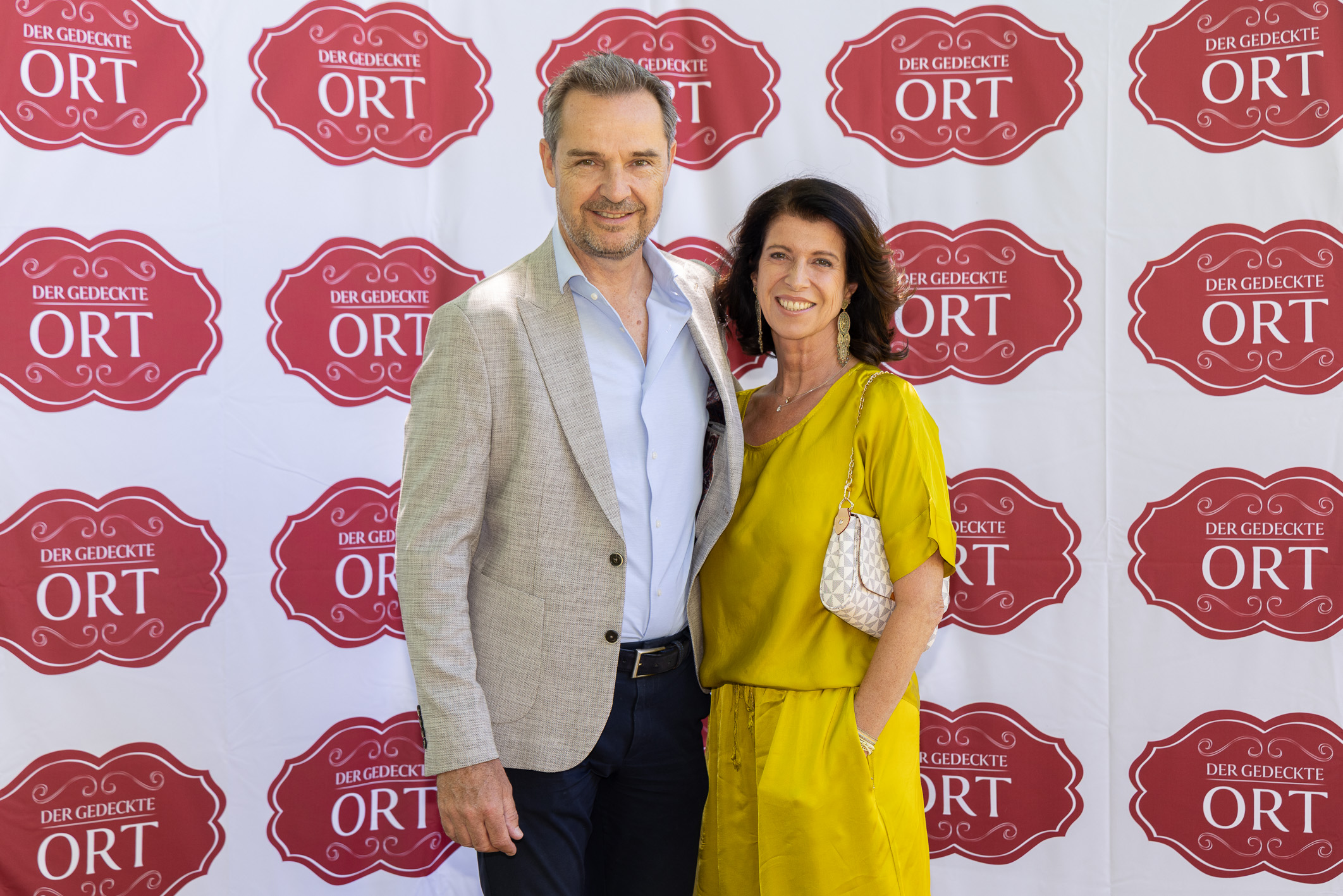 Ein Mann in einem grauen Blazer und eine Frau in einem gelben Kleid stehen lächelnd vor einer rot-weißen Veranstaltungskulisse mit der Aufschrift „Der gedeckte Ort“.