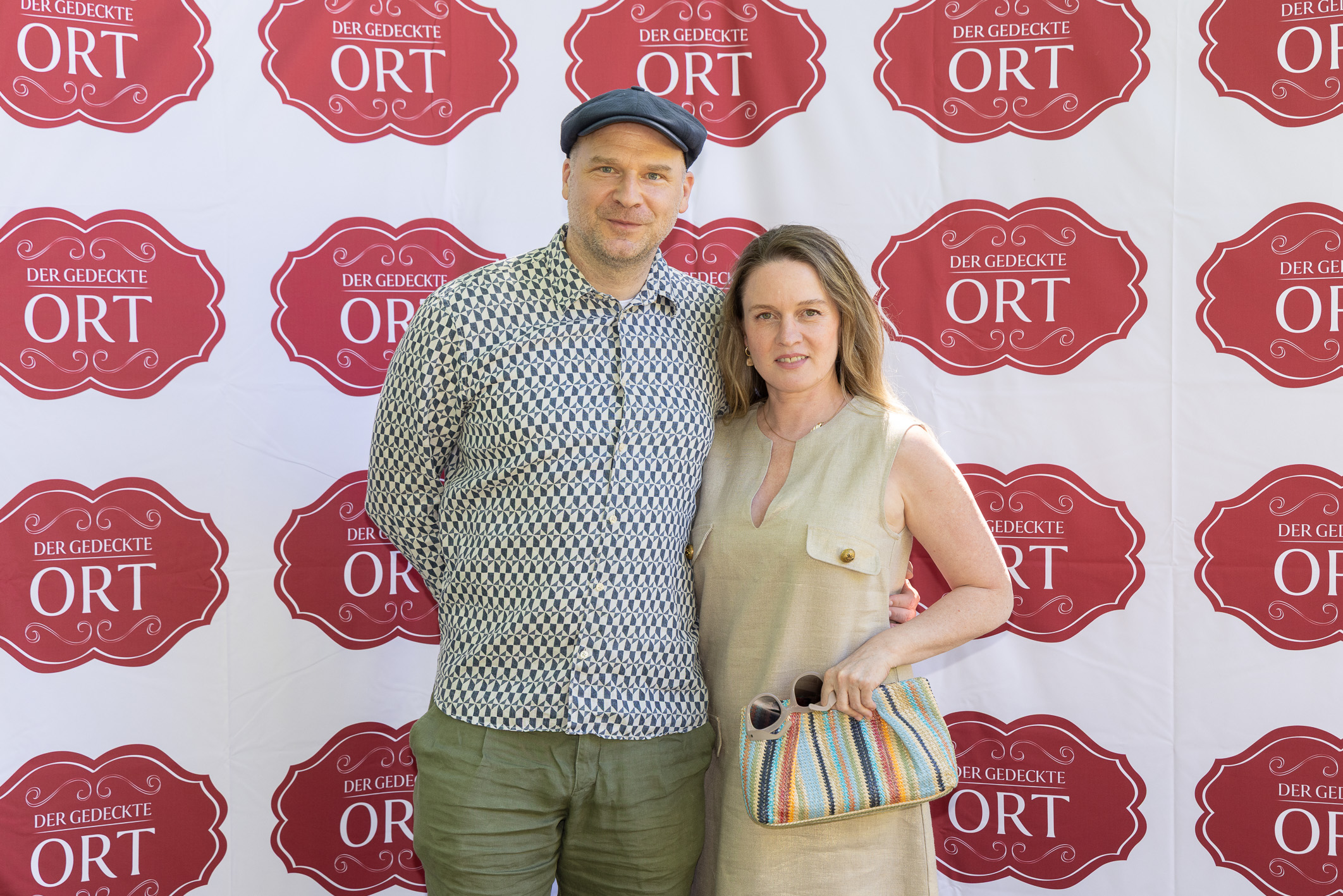 Ein Mann und eine Frau posieren gemeinsam vor einem Hintergrund mit roten „Der gedeckte ORT“-Logos. Die Frau hält eine bunt gestreifte Tasche in der Hand.