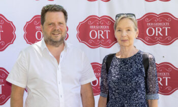 Ein Mann in einem weißen Hemd und eine Frau in einem blauen Kleid stehen vor einer Veranstaltungskulisse mit der Aufschrift „Der Gedeckte Ort“.