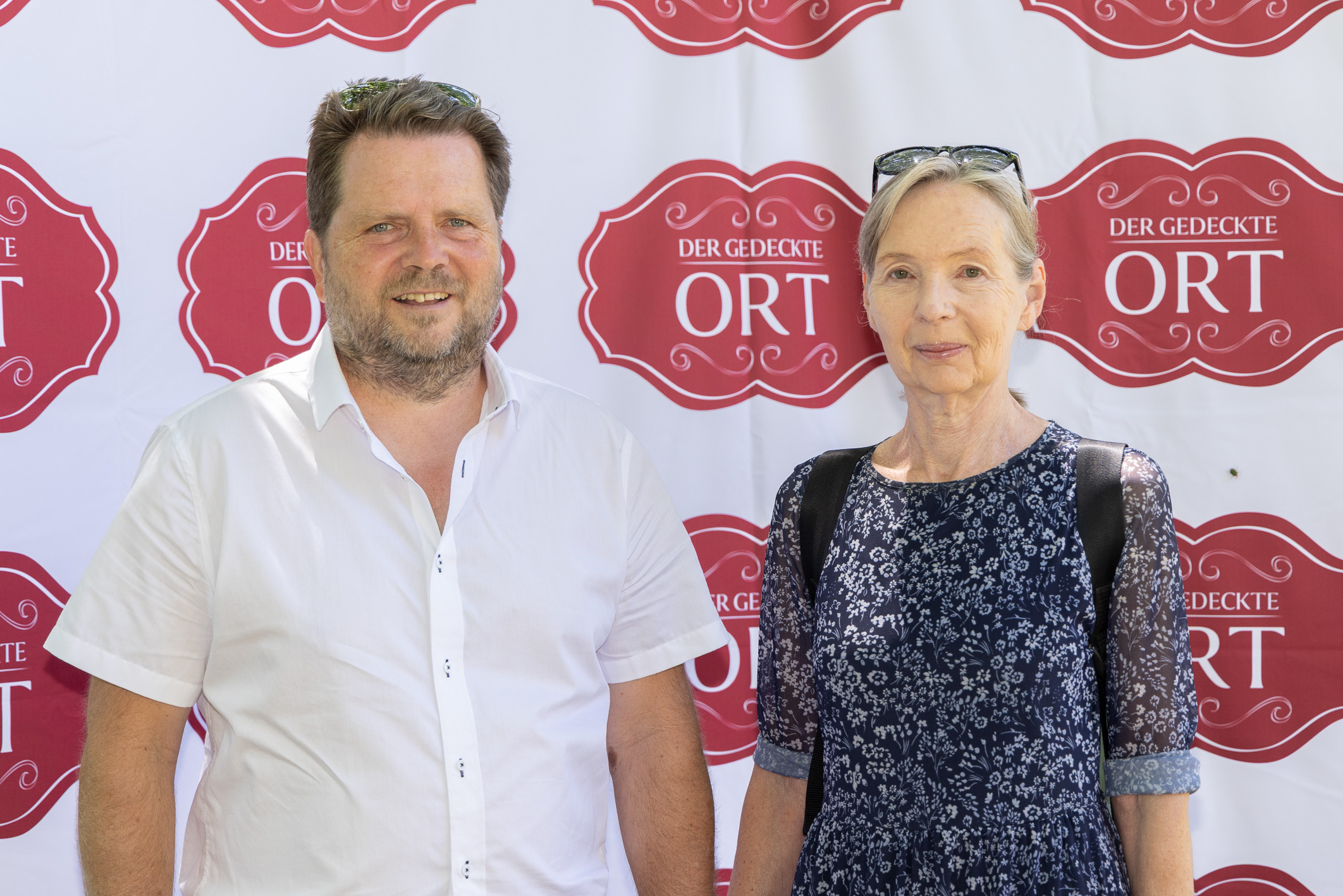 Ein Mann in einem weißen Hemd und eine Frau in einem blauen Kleid stehen vor einer Veranstaltungskulisse mit der Aufschrift „Der Gedeckte Ort“.