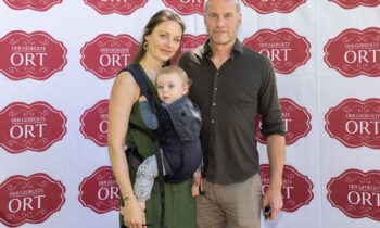 Eine Frau, ein Mann und ein Baby posieren gemeinsam vor einem Hintergrund, auf dem das rote „Der gedeckte Ort“-Logo wiederholt abgebildet ist.
