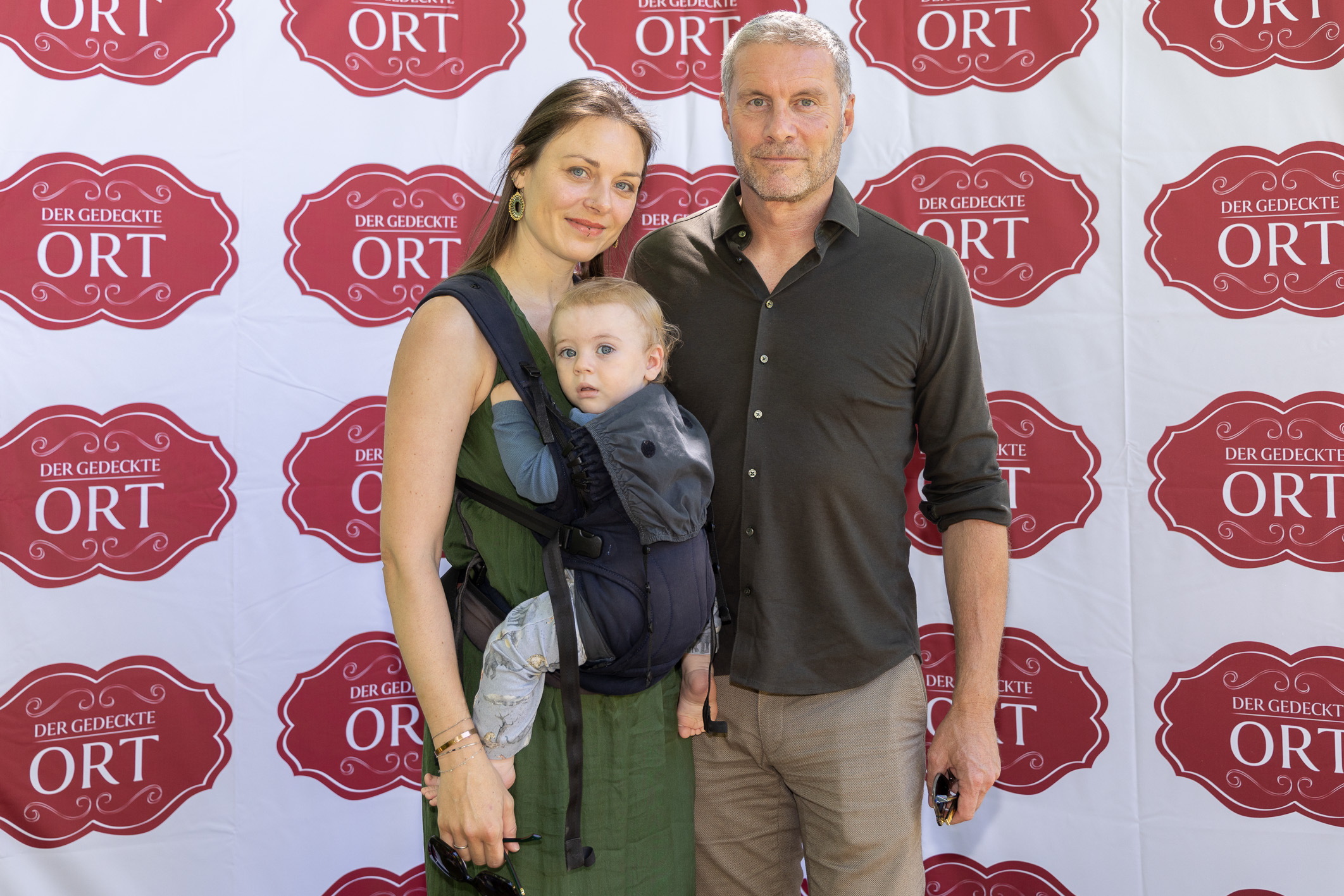Eine Frau, ein Mann und ein Baby posieren gemeinsam vor einem Hintergrund, auf dem das rote „Der gedeckte Ort“-Logo wiederholt abgebildet ist.
