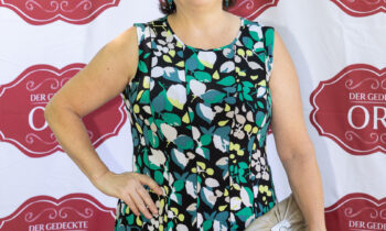 Eine Frau in einem ärmellosen, gemusterten Kleid lächelt mit einer Clutch in der Hand vor einem „Der Gedeckte Ort Vol. 2“-Step-and-Repeat-Banner.