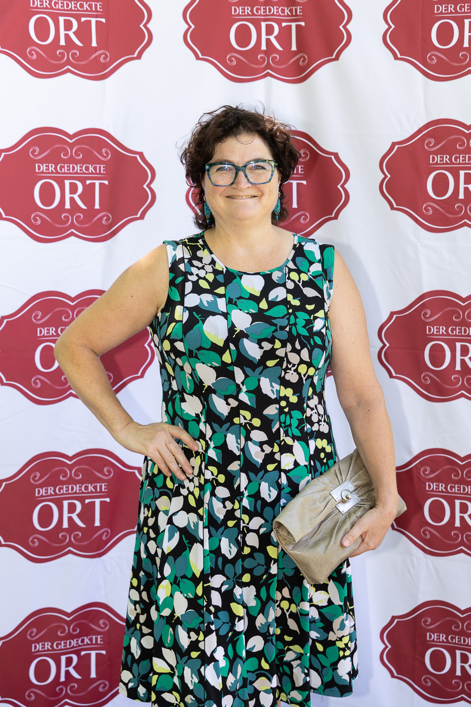 Eine Frau in einem ärmellosen, gemusterten Kleid lächelt mit einer Clutch in der Hand vor einem „Der Gedeckte Ort Vol. 2“-Step-and-Repeat-Banner.