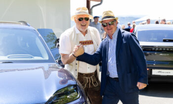 Zwei lächelnde Männer, einer in traditioneller Tracht und einer im Anzug, reichen sich an einem sonnigen Tag neben geparkten Autos die Hände – auf dem Cover von „Der gedeckte Ort, Band 2“.
