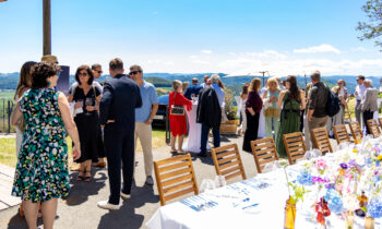 Eine Gruppe von Menschen, die sich im Freien an einem langen, für ein Essen gedeckten Tisch mit Blumen und Gläsern unterhält, vor dem Hintergrund eines sonnigen blauen Himmels und einer malerischen Landschaft.