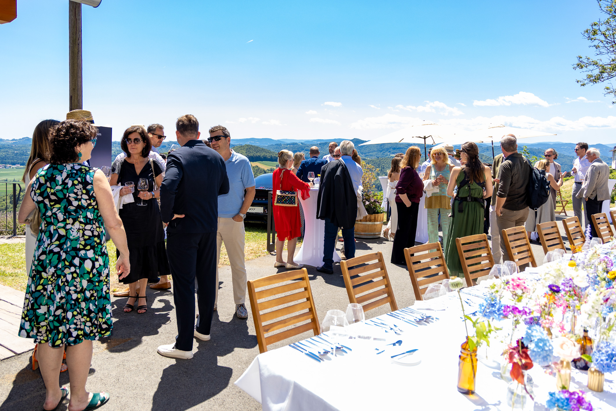 Eine Gruppe von Menschen, die sich im Freien an einem langen, für ein Essen gedeckten Tisch mit Blumen und Gläsern unterhält, vor dem Hintergrund eines sonnigen blauen Himmels und einer malerischen Landschaft.