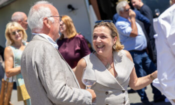 Zwei Menschen, die bei einer Veranstaltung im Freien lächeln und sich unterhalten, während sie Weingläser in den Händen halten; im Hintergrund unterhalten sich andere Gäste miteinander.