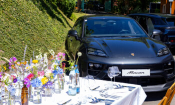 Ein schwarzer Porsche Macan steht in der Nähe eines Tisches, der für ein Essen im Freien gedeckt ist – mit bunten Blumen, Gläsern und Tellern.