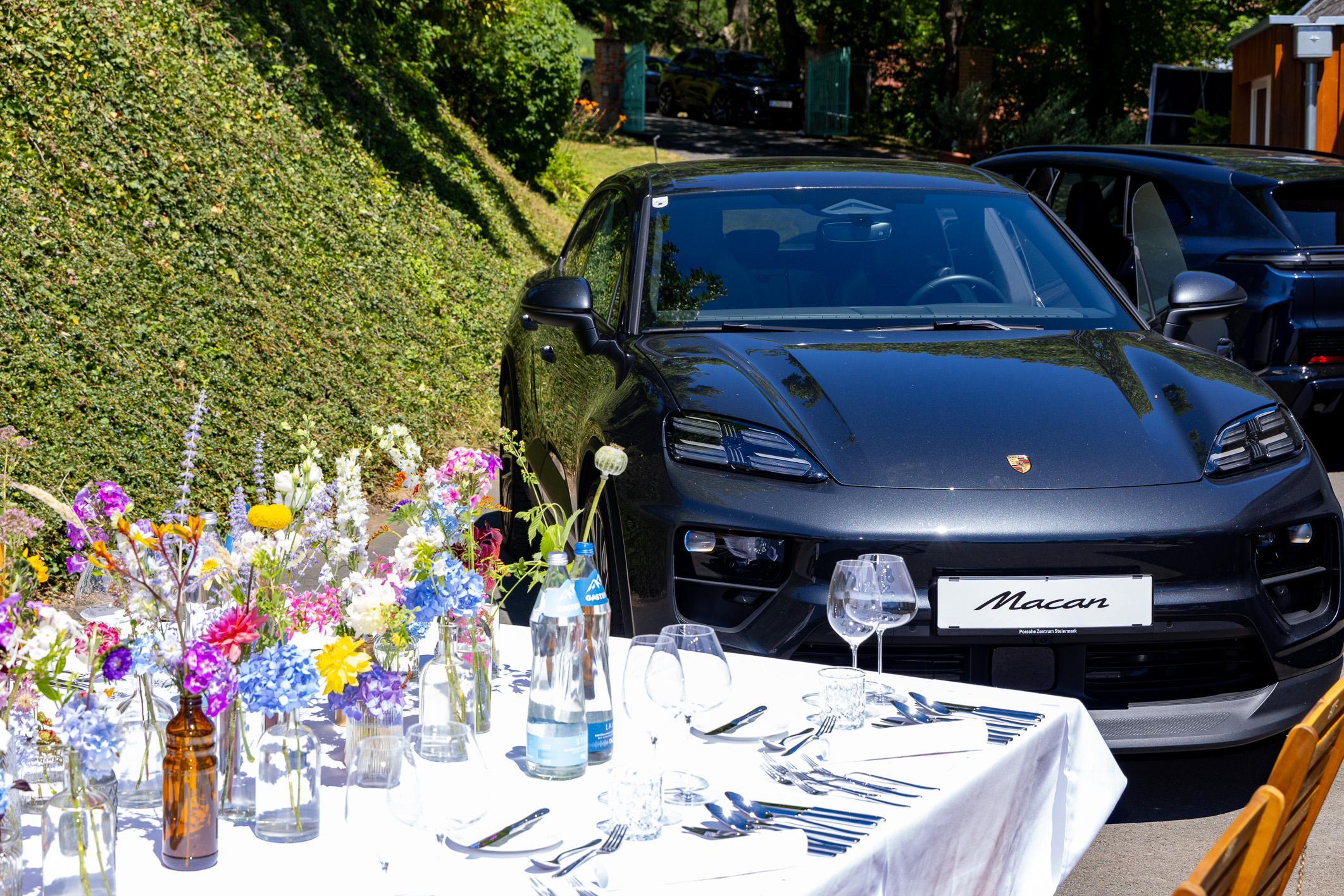 Ein schwarzer Porsche Macan steht in der Nähe eines Tisches, der für ein Essen im Freien gedeckt ist – mit bunten Blumen, Gläsern und Tellern.