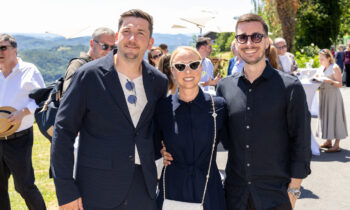 Drei Personen lächeln und posieren gemeinsam im Freien bei einer sonnigen Veranstaltung, im Hintergrund sind weitere Gäste und Grünflächen zu sehen.