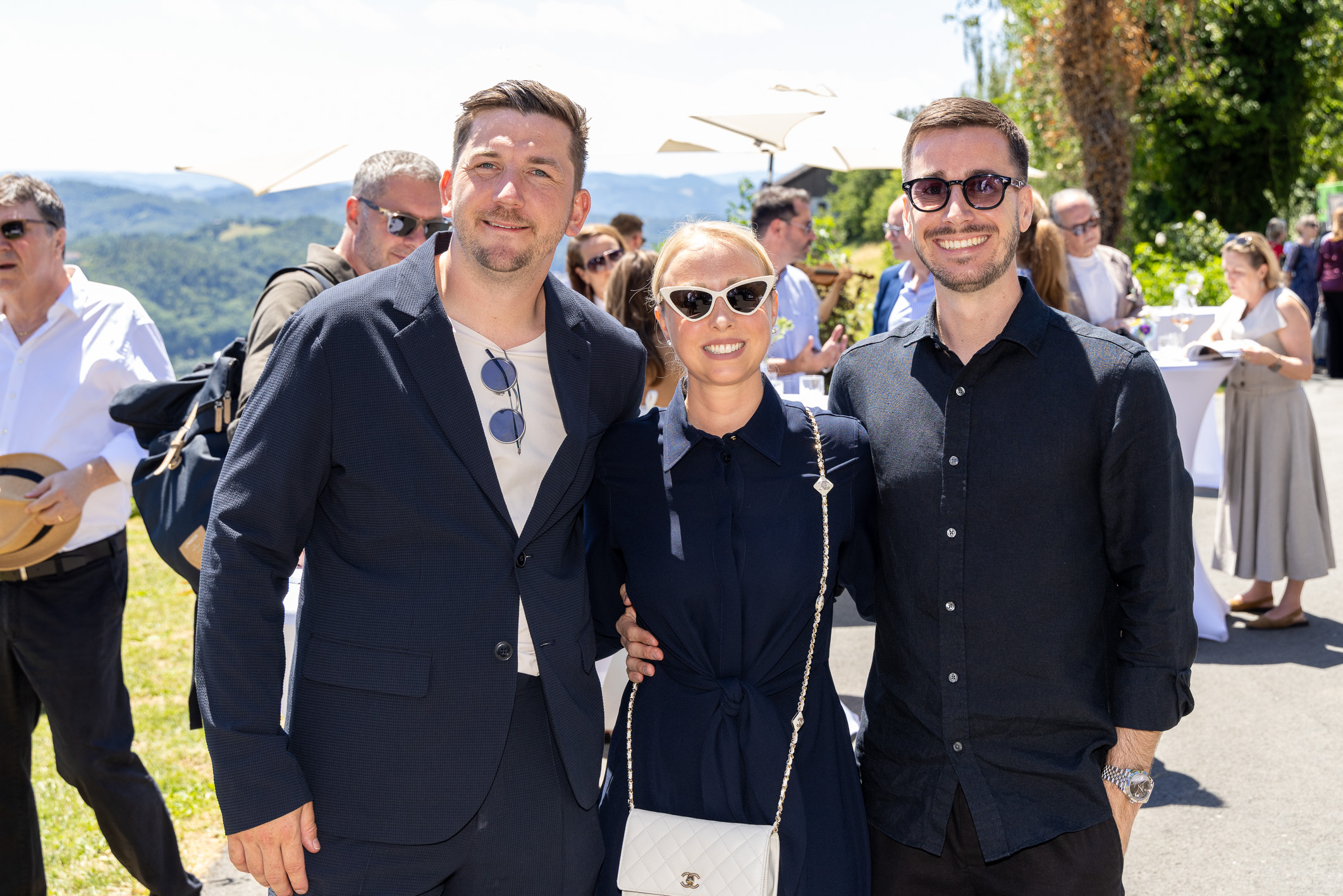 Drei Personen lächeln und posieren gemeinsam im Freien bei einer sonnigen Veranstaltung, im Hintergrund sind weitere Gäste und Grünflächen zu sehen.