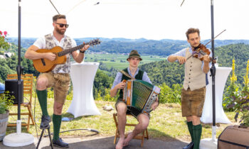 Drei Männer in traditioneller bayerischer Tracht spielen im Freien Gitarre, Akkordeon und Geige, vor dem Hintergrund malerischer Hügel und grüner Landschaft.