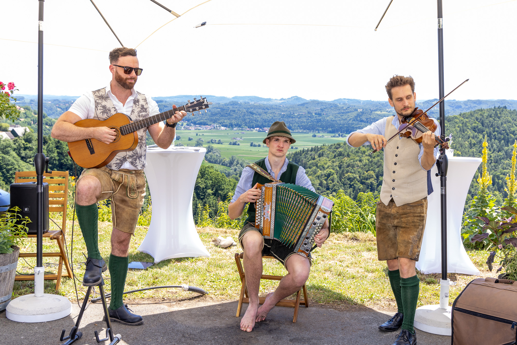 Drei Männer in traditioneller bayerischer Tracht spielen im Freien Gitarre, Akkordeon und Geige, vor dem Hintergrund malerischer Hügel und grüner Landschaft.