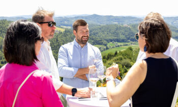 Vier Menschen stehen draußen um einen Tisch herum, unterhalten sich und lächeln, im Hintergrund sind malerische grüne Hügel und strahlender Sonnenschein zu sehen.